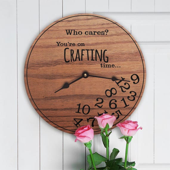 A wooden circle clock is hung on a white wooden wall, with pink roses touching the bottom in the foreground. The clock has two black hands showing the time is 8:16. The clock numbers are all in the bottom of the clock as if they fell down. The text on top of the clock says "Who cares? You're on crafting time..."