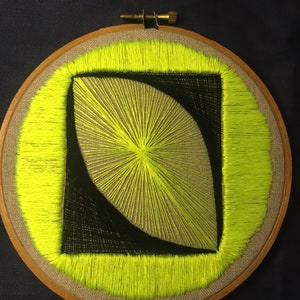 May include: A circular embroidery hoop with a geometric design. The design is made with black and neon yellow thread. The design is a curved shape with a radiating pattern.