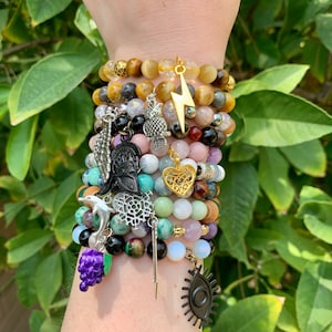 May include: A stack of bracelets with various gemstone beads and charms, including a heart, a lightning bolt, a dolphin, a grape, and an eye. The bracelets are on a person's wrist.