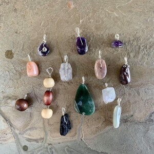 Puede incluir: Una colección de 14 colgantes únicos con diversas piedras y materiales, incluyendo amatista, ágata, madera y obsidiana. Cada colgante está hecho a mano con alambre envuelto y está listo para ser añadido a un collar o pulsera.