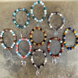 Pode incluir: Dez pulseiras de contas com vários pingentes, incluindo um coração, uma cruz, uma engrenagem, um camelo, um elefante e um lagarto. As pulseiras são feitas com pedras de diferentes cores, incluindo azul, verde, marrom, vermelho e preto.