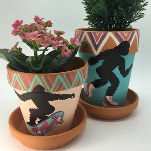May include: Two terracotta flower pots painted with a whimsical design. One pot features a silhouette of a Bigfoot skateboarding on a pink and purple skateboard. The other pot features a silhouette of a Bigfoot ice skating on a turquoise background. Both pots have a geometric pattern around the top.