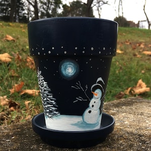 Könnte beinhalten: Dunkelblau bemalter Blumentopf mit passender Untertasse. Der Topf zeigt eine Winterszene mit einem Schneemann, einem Vollmond, schneebedecktem Boden und einem Nadelbaum. Weiße Punkte stellen Sterne dar.