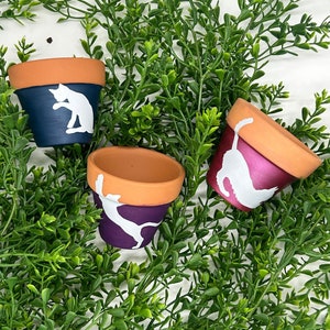 May include: Three small terracotta flower pots with white painted cat silhouettes. The pots are painted in shades of blue, purple, and pink.