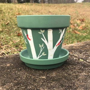 Könnte beinhalten: Ein grüner Terrakotta-Blumentopf mit einem bemalten Design aus weißen Birken und roten Kardinälen. Der Topf hat eine passende Untertasse.