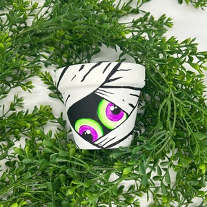 Puede incluir: Una pequeña maceta pintada de blanco decorada con una cara de momia. Los ojos de la momia son morados y verdes, y la maceta está rodeada de follaje verde. La maceta es una decoración con temática de Halloween.