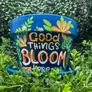 Puede incluir: Una maceta pintada de azul con las palabras "Good Things Bloom Here" en blanco, naranja y amarillo. La maceta está decorada con flores naranjas y blancas y hojas verdes. La maceta se asienta sobre un platillo azul a juego.