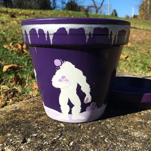 May include: A purple painted flower pot with a white Bigfoot design. The pot has a silver drip design around the rim and a matching saucer. The Bigfoot is holding a cluster of purple flowers.