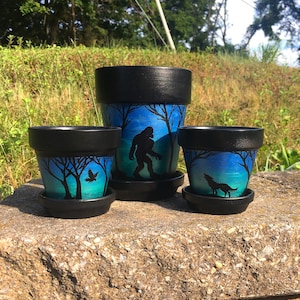 May include: Three black terracotta plant pots with blue and black painted designs. The largest pot features a silhouette of Bigfoot in a forest scene. The medium pot features a silhouette of a bird in a forest scene. The smallest pot features a silhouette of a wolf in a forest scene.
