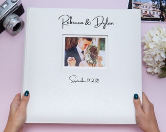 May include: A white wedding photo album with a rectangular photo window. The album cover has the names "Rebecca & Dylan" and the date "September 19, 2021" printed in black script. The album is held by two hands.