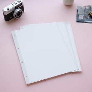May include: A collection of white photo album pages with three-hole punches, stacked on a pink surface. A vintage black and silver camera sits nearby, along with a stack of printed photos and a gold pen. A small white vase is also present.