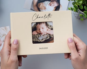 May include: A cream-colored photo album with a window showing a baby sleeping in a basket. The album has the name "Charlotte" and the date "04.03.2025" printed on the front.