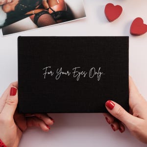 May include: A black box with the text "For Your Eyes Only" in cursive script. The box is held by two hands with red nail polish. A photograph and two red heart-shaped decorations are in the background.