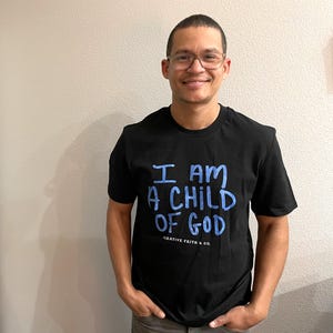 May include: Black crew neck t-shirt with the text "I AM A CHILD OF GOD" in blue. The shirt also features the text "CREATIVE FAITH & CO." in a smaller font. The person is wearing glasses and has their hands in their pockets.