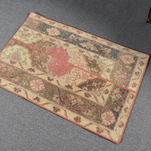 May include: Rectangular rug with a faded, vintage appearance. The rug features a central panel in a muted red hue, surrounded by floral and geometric patterns in shades of green, beige, and dark blue. The border is a light orange.