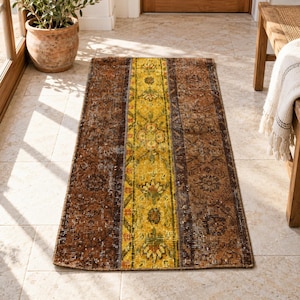 Könnte beinhalten: Ein rechteckiger Teppich mit vertikalem Patchwork-Design. Das mittlere Feld ist in leuchtendem Gelb gehalten und mit komplexen Blumenmustern in Grün und Orange verziert, flankiert von zwei breiteren Feldern in Dunkelbraun und Rost mit Blumenmotiven.