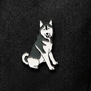 May include: A black and white enamel pin shaped like a sitting husky dog with its tongue sticking out, attached to a black fabric surface with a visible zipper and a black cord pull tab at the top corner of the textured material.