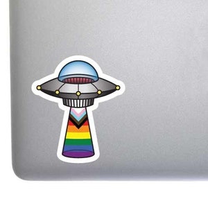 May include: A sticker of a cartoon UFO with a rainbow and transgender flag design. The UFO is silver with a blue dome and red interior. The sticker is placed on a gray laptop.