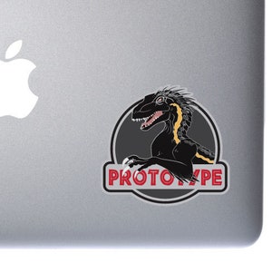 May include: A silver laptop with a white Apple logo and a black and yellow dinosaur sticker with the text "PROTOTYPE" in red.