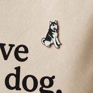 May include: A beige canvas tote bag featuring the black printed text "I love my dog." and a small enamel pin of a black and white husky dog attached to the front. The bag has two matching fabric handles and a simple, structured rectangular shape.