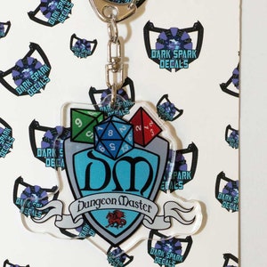 May include: A clear acrylic keychain featuring a blue shield with the letters "DM" for Dungeon Master, a dragon, and a banner with the words "Dungeon Master".  Four colorful dice are attached to the shield.
