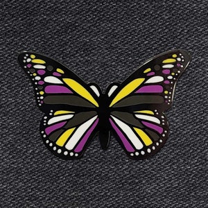 May include: A butterfly-shaped enamel pin with a non-binary pride flag design. The pin features a black body and wings with yellow, purple, white, and black sections. The pin is set against a dark gray denim background.