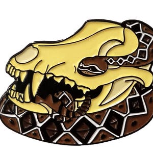 May include: A yellow enamel pin of a snake wrapped around a skull. The snake is brown and black with a diamond pattern. The skull has sharp teeth and is set against a brown background.