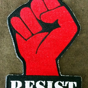 May include: Red fabric patch with a raised fist and the word "RESIST" in white letters on a black background.