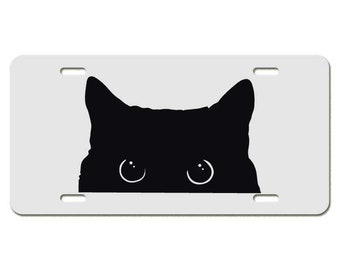 Puede incluir: Una silueta de gato negro con ojos grandes mirando por encima de una placa de matrícula blanca.