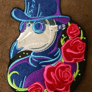 May include: Embroidered patch featuring a stylized plague doctor wearing a blue top hat and a white mask with a long beak. The doctor is surrounded by red roses with green leaves on a black background.