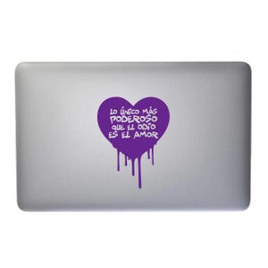 May include: A silver laptop with a purple heart-shaped sticker. The sticker has the Spanish text "Lo único más poderoso que el odio es el amor" in white, with dripping paint effect. The sticker is centered on the laptop's lid.