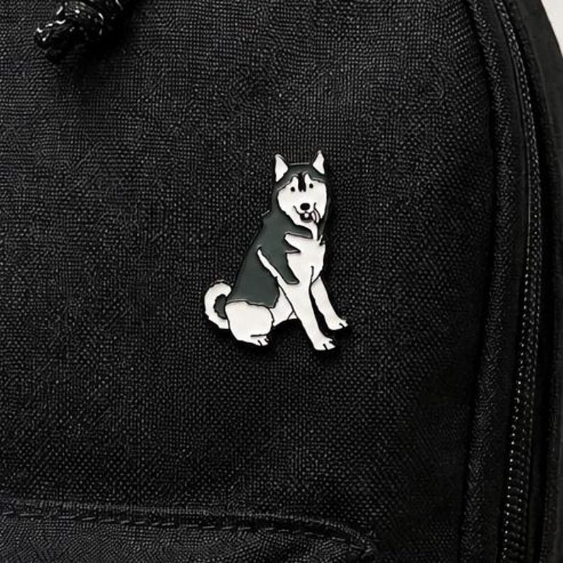 May include: A black and white enamel pin shaped like a sitting husky dog with its tongue sticking out, attached to a black fabric surface with a visible zipper and a black cord pull tab at the top corner of the textured material.