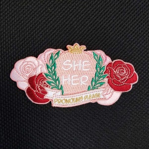 May include: Pink and red embroidered patch with a crown, roses, and the text "She Her Pronouns Please".