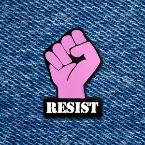 May include: Pink raised fist with a black outline and the word "RESIST" in white letters on a black background.