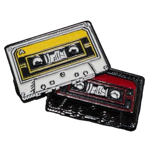 May include: Two vintage cassette tapes, one white with a yellow label and one black with a red label. Both tapes have a silver metal finish and are stacked on top of each other.