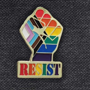 May include: A gold-colored enamel pin with a raised fist in the colors of the Progress Pride flag. The fist is raised in the air and the word "RESIST" is written in rainbow colors below the fist.