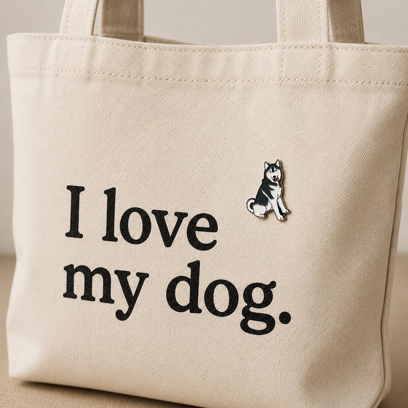 May include: A beige canvas tote bag featuring the black printed text "I love my dog." and a small enamel pin of a black and white husky dog attached to the front. The bag has two matching fabric handles and a simple, structured rectangular shape.