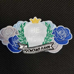 May include: White embroidered patch with a crown, a tombstone with the text "HE/HIM" and a banner that reads "PRONOUNS PLEASE". The patch is surrounded by three embroidered roses in shades of blue, white, and gray.