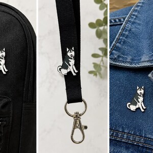 May include: A black and white enamel pin shaped like a sitting Siberian Husky dog. The pin is shown attached to a black backpack, a black fabric lanyard with a silver metal lobster clasp, and the collar of a blue denim jacket.