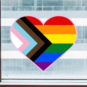 May include: A heart-shaped sticker with a rainbow flag design, incorporating the colors of the transgender pride flag and the black and brown stripes of the progress pride flag.