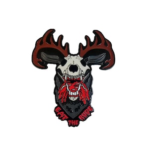 May include: A black and red enamel pin featuring a skull with antlers and a bloody mouth. The text "Eat the Ride" is written in a banner below the skull.