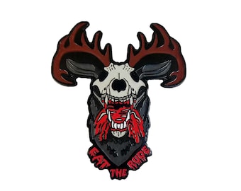 May include: A black and red enamel pin featuring a skull with antlers and a bloody mouth. The text "Eat the Ride" is written in a banner below the skull.