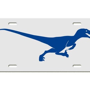 May include: A white license plate featuring a blue silhouette of a running dinosaur. The dinosaur is a raptor, with its mouth open and tail extended. The plate has four mounting holes.