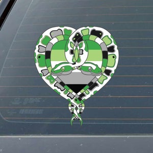 May include: A heart-shaped sticker featuring two green snakes intertwined. The snakes have green, black, and gray patterns, with the AroAce pride flag colors incorporated. The design is set against a window.