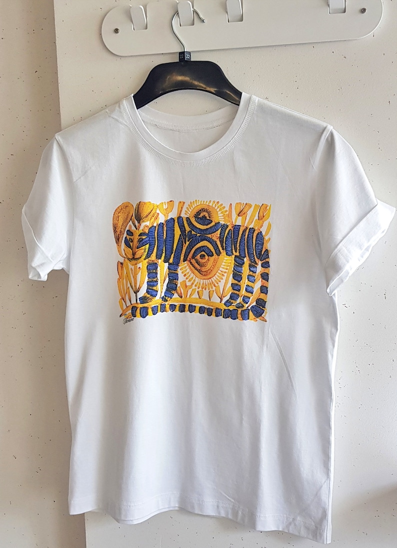 Op de afbeelding: Wit T-shirt met een blauwe en gele grafische print van een gestileerde tijger. De tijger is omringd door gele bloemen en zonnen.