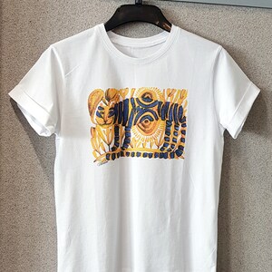 Op de afbeelding: Wit T-shirt met een kleurrijke afbeelding van een tijger met gele, blauwe en oranje details. De tijger is omringd door gestileerde bloemen en bladeren.