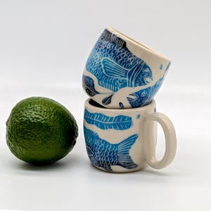 May include: Two white ceramic mugs with blue fish designs. The mugs are stacked on top of each other. The top mug features a fish with its mouth open, and the bottom mug has a fish with its tail fin visible.