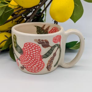Könnte beinhalten: Weiße Keramiktasse mit Blumenmuster. Das Design zeigt rote und grüne Blumen mit weißen Punkten.