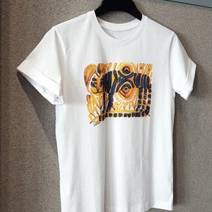 Op de afbeelding: Wit T-shirt met een kleurrijke grafische print van een tijger met blauwe en gele strepen en een zon-achtig ontwerp. De grafiek is omgeven door gele en blauwe bloemmotieven.