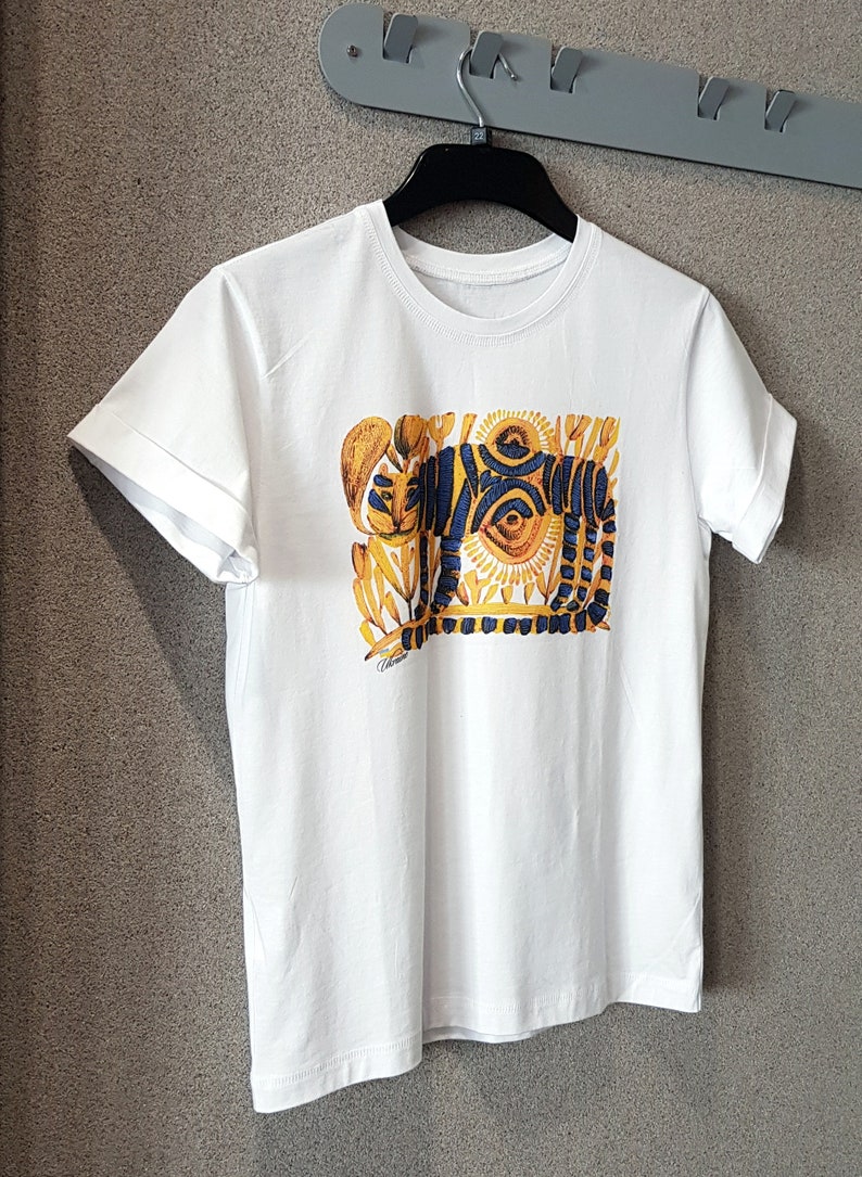 Op de afbeelding: Wit T-shirt met een kleurrijke grafische print van een tijger met blauwe en gele strepen en een zon-achtig ontwerp. De grafiek is omgeven door gele en blauwe bloemmotieven.